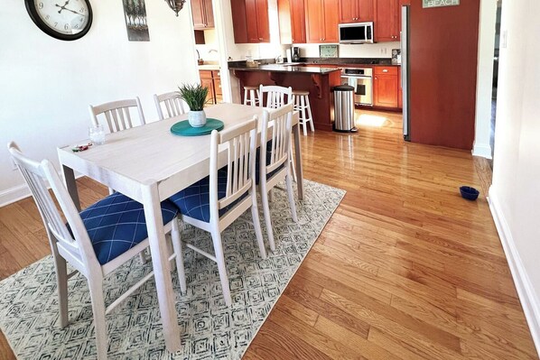 A dining area with a white table and chairs, a patterned rug, and a kitchen in the background.