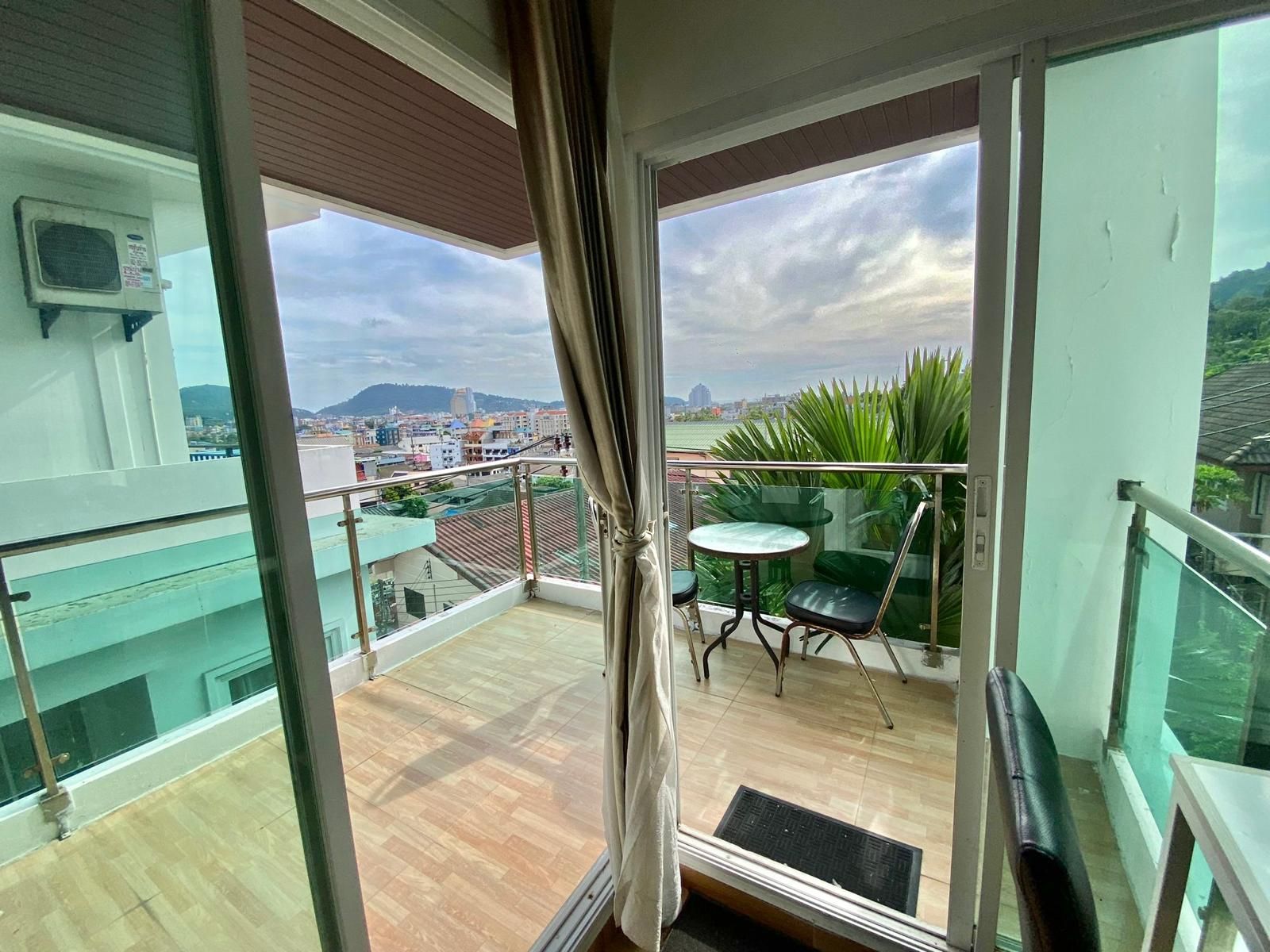 Ein Balkon mit Glastür, einem kleinen Tisch und einem Stuhl, mit Blick auf eine Stadtlandschaft mit Gebäuden und Grünflächen.
