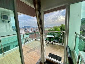 Un balcó amb una porta de vidre, una taula petita i una cadira, amb vista a un paisatge urbà ple d'edificis i vegetació.