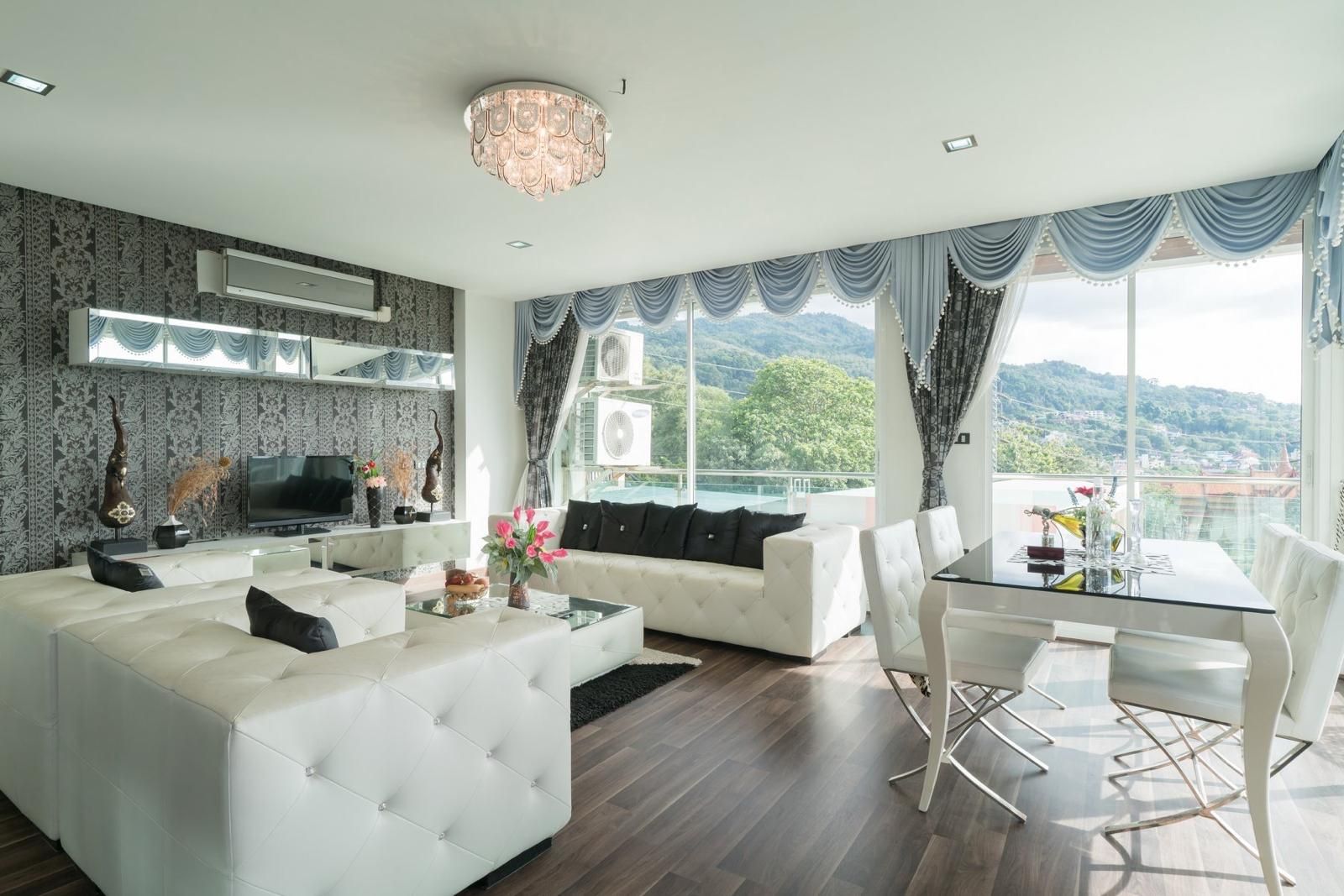 A modern living room with a sofa, a coffee table, and a dining area with chairs. There is a TV on a stand, decorative items, and a view of the outdoors.