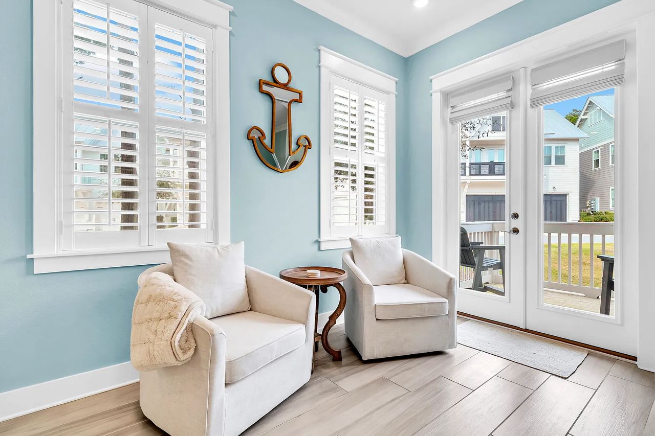 A room with a light blue wall, two white chairs, a small round table, and a large window with shutters.