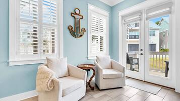 A room with a light blue wall, two white chairs, a small round table, and a large window with shutters.