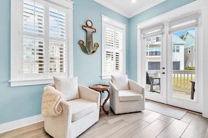 A room with a light blue wall, two white chairs, a small round table, and a large window with shutters.