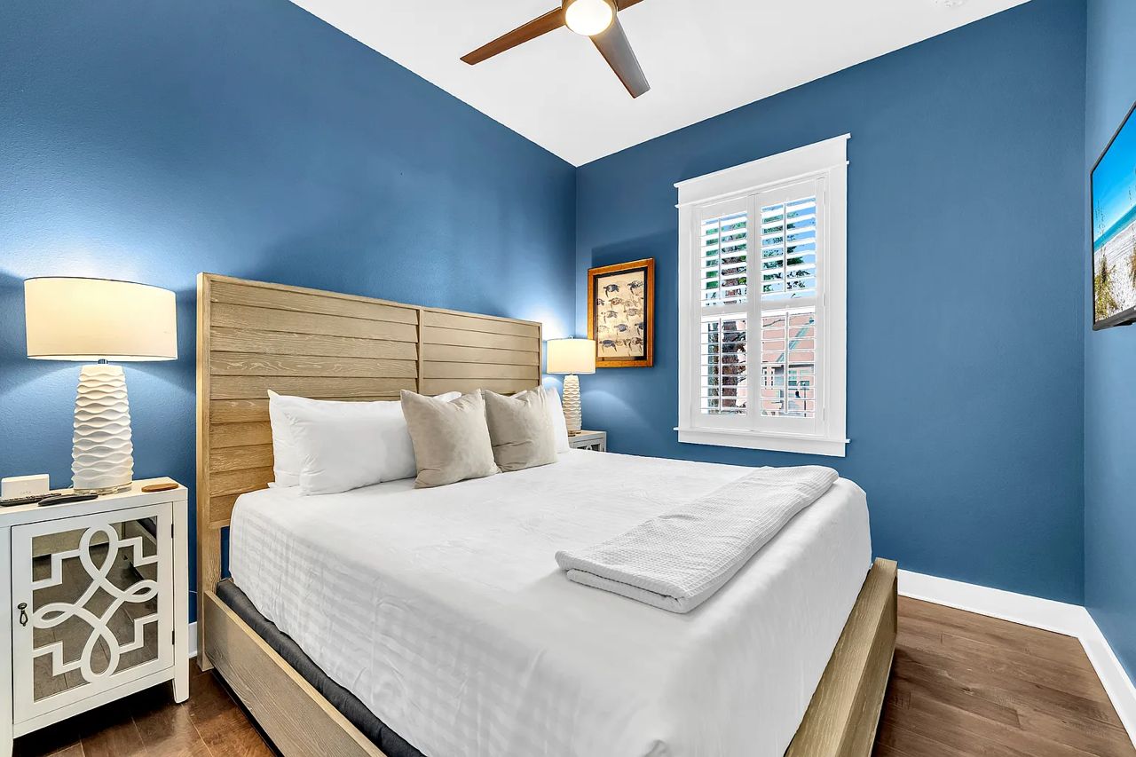 A bedroom with a bed, two bedside tables, a ceiling fan, a window with blinds, and a wall-mounted picture.