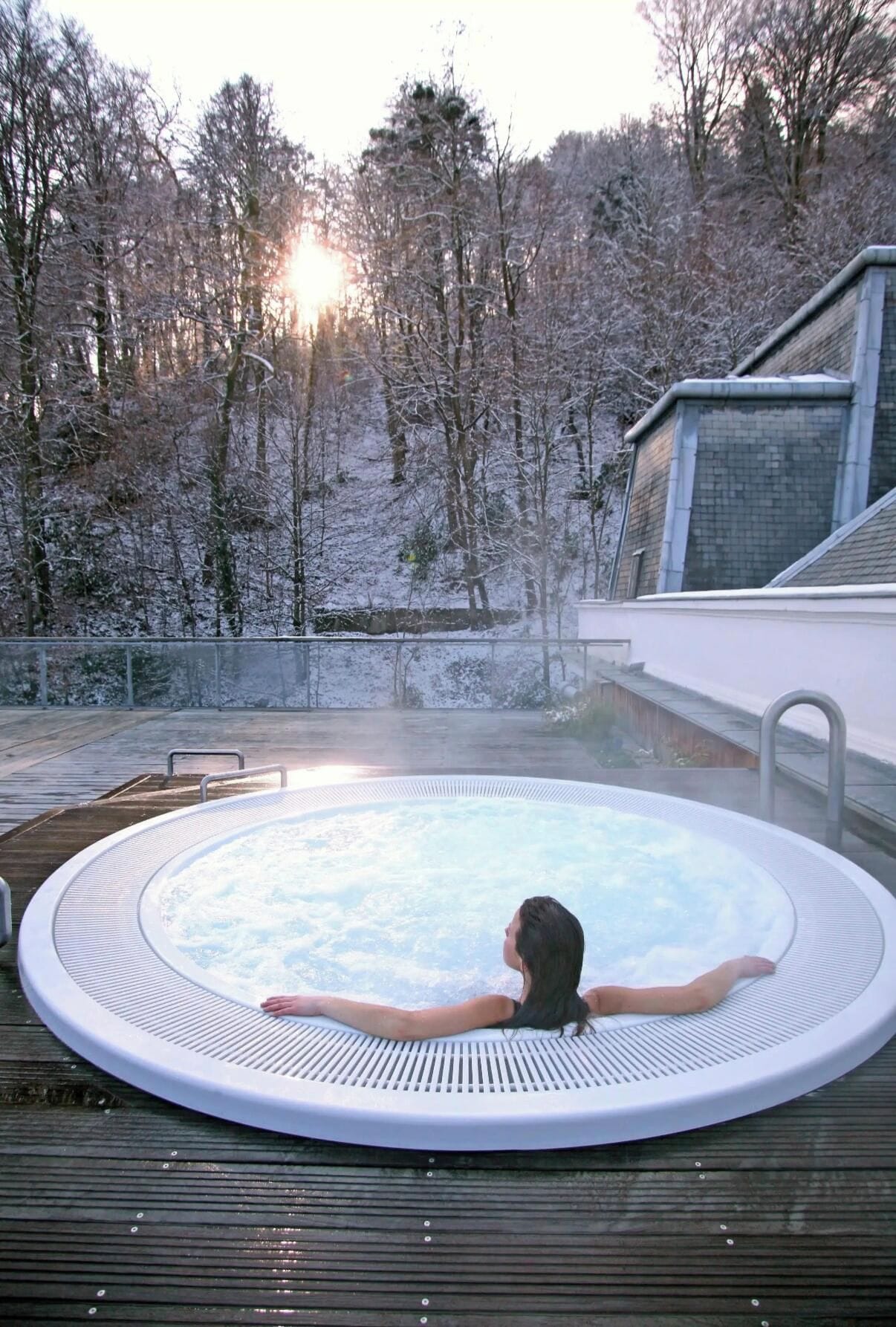 A person relaxing in a hot tub outdoors with a view of a snowy forest and a building in the background.