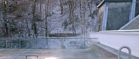 Une personne se détend dans un bain à remous en plein air, avec vue sur une forêt enneigée et un bâtiment en arrière-plan.