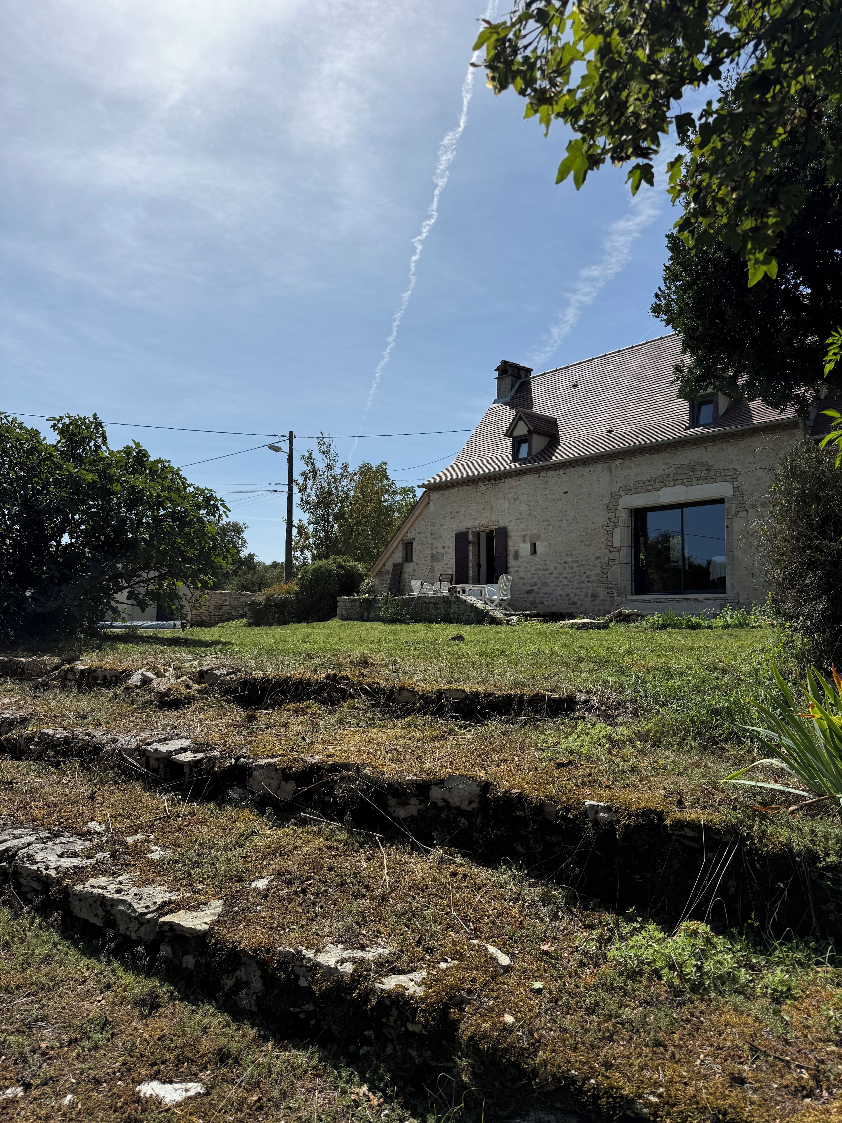 Une maison en pierre avec un toit en pente, une allée pavée et une pelouse bordée de murets en pierre.