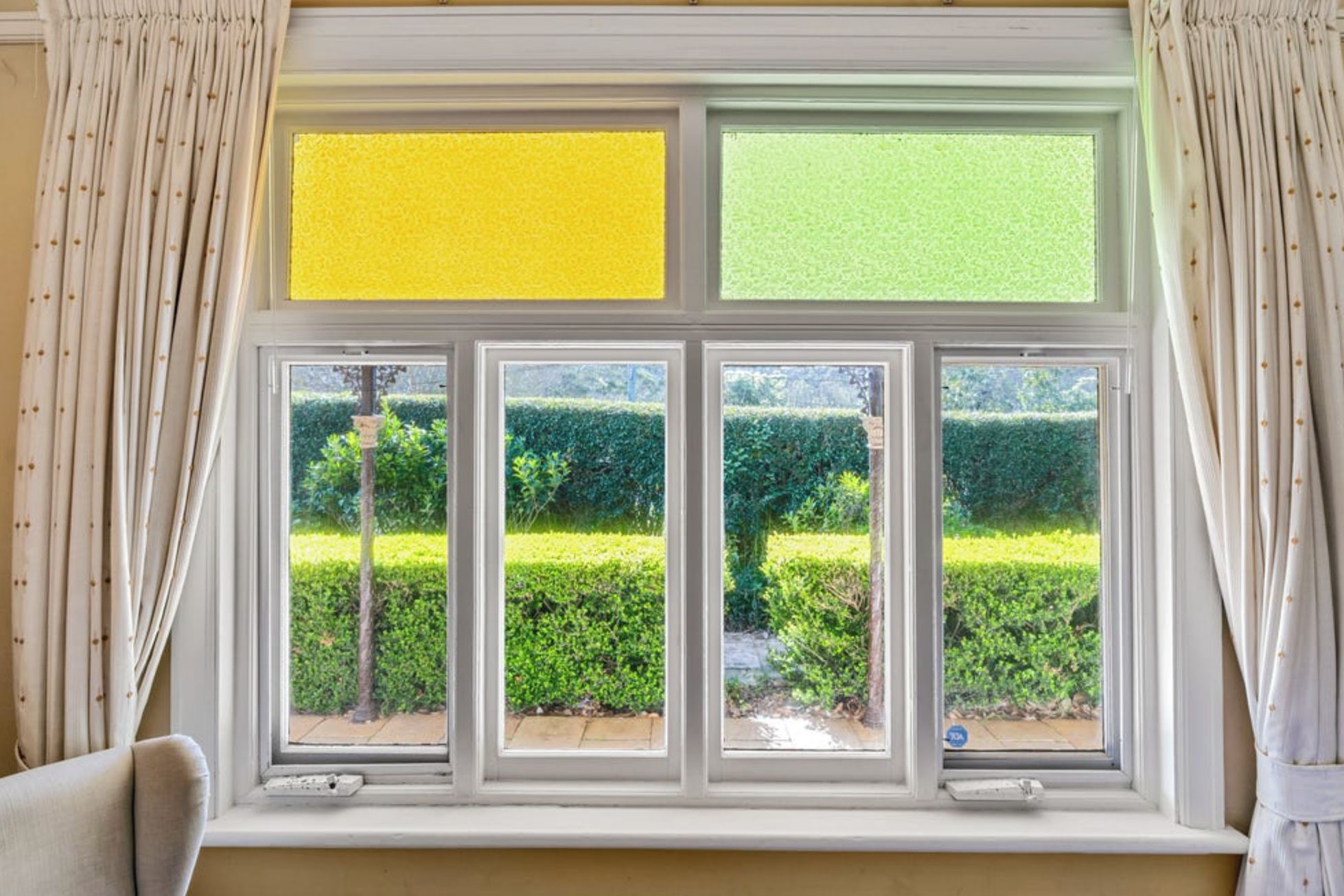 A window with a view of a neatly trimmed hedge and a tree, curtains on either side, and a thermostat on the window sill.