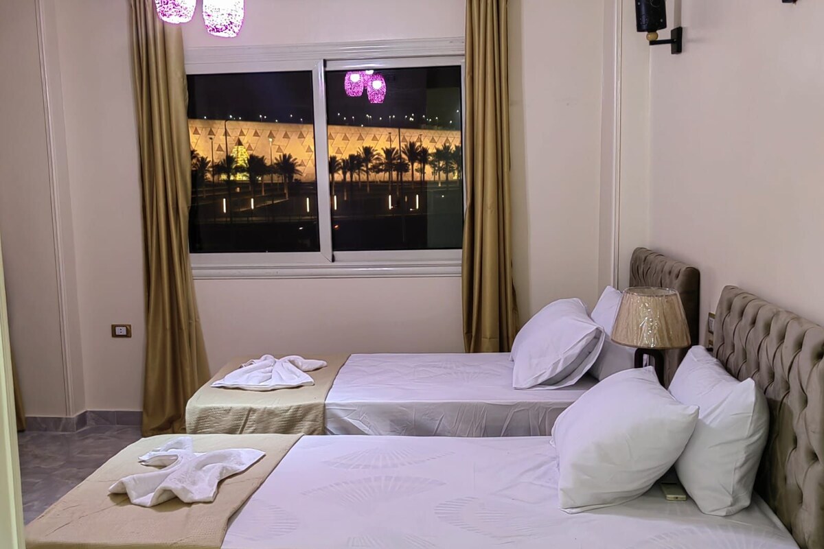 A hotel room with two beds, a nightstand, a lamp, and a window with a view of a building and palm trees.