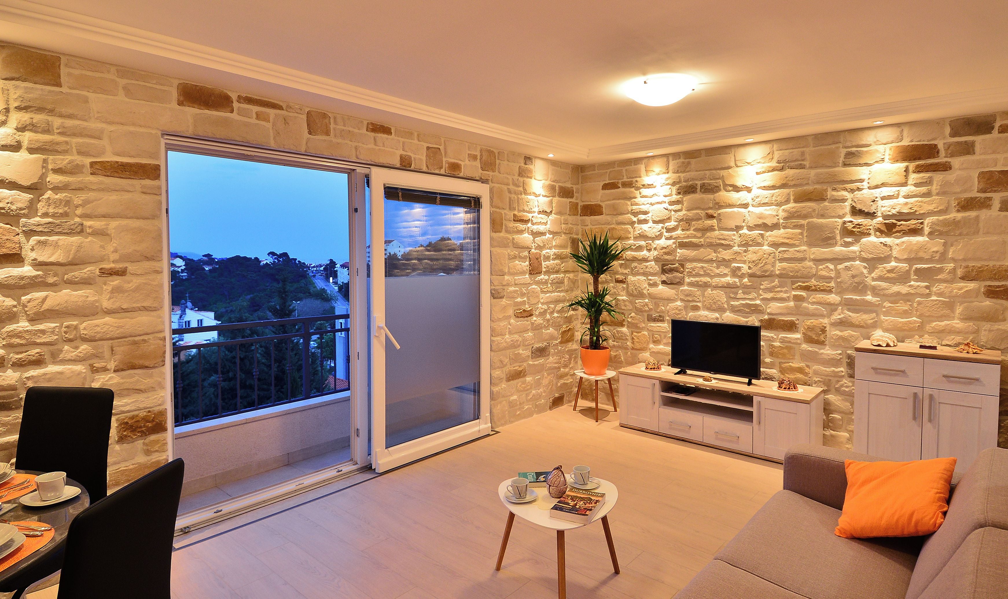 A modern living room with a stone wall, a sliding glass door to a balcony, a flat-screen TV, and a sofa with an orange pillow.
