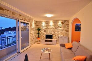 A living room with a stone wall, a television, a sofa, and a balcony with a view of buildings. - Welcoming 1-bedroom apartment with AC in Dubrovnik with private parking place (Dubrovnik)