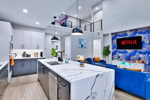 A modern kitchen with a central island, a dining area with blue sofas, and a living room with a Netflix sign. - Luxe Living in the heart of Dallas (Dallas)