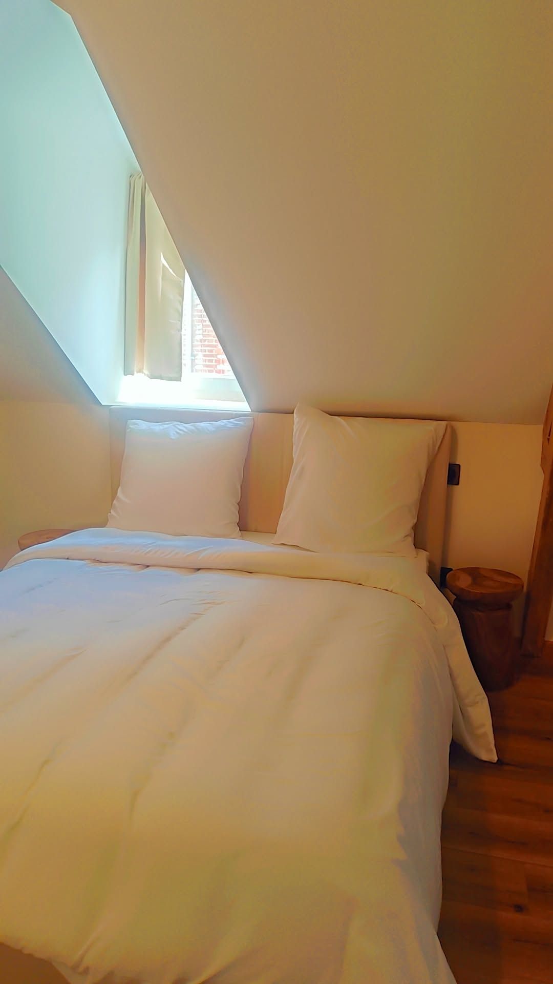A bedroom with a sloped ceiling, a bed with white bedding, a bedside table, and a small wooden stool.