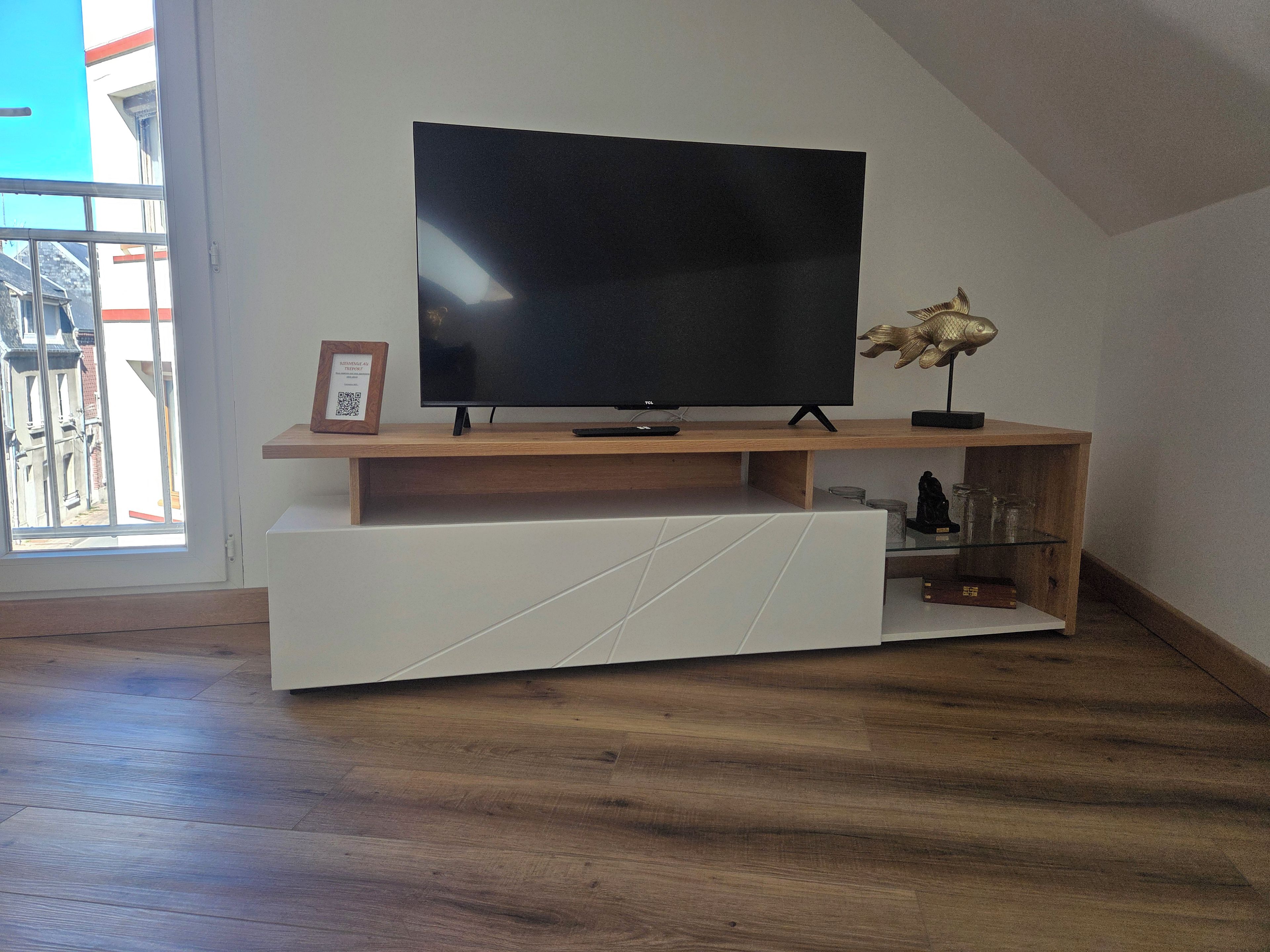 A modern living room with a flat-screen TV on a wooden stand, a white entertainment unit, and a glass shelf with decorative items.