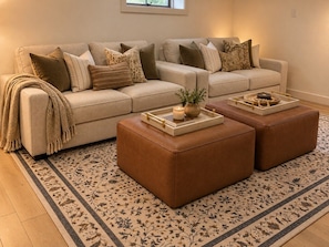 Un soggiorno con un divano beige, due poggiapiedi in pelle, un tappeto decorato e un tavolino con oggetti ornamentali.