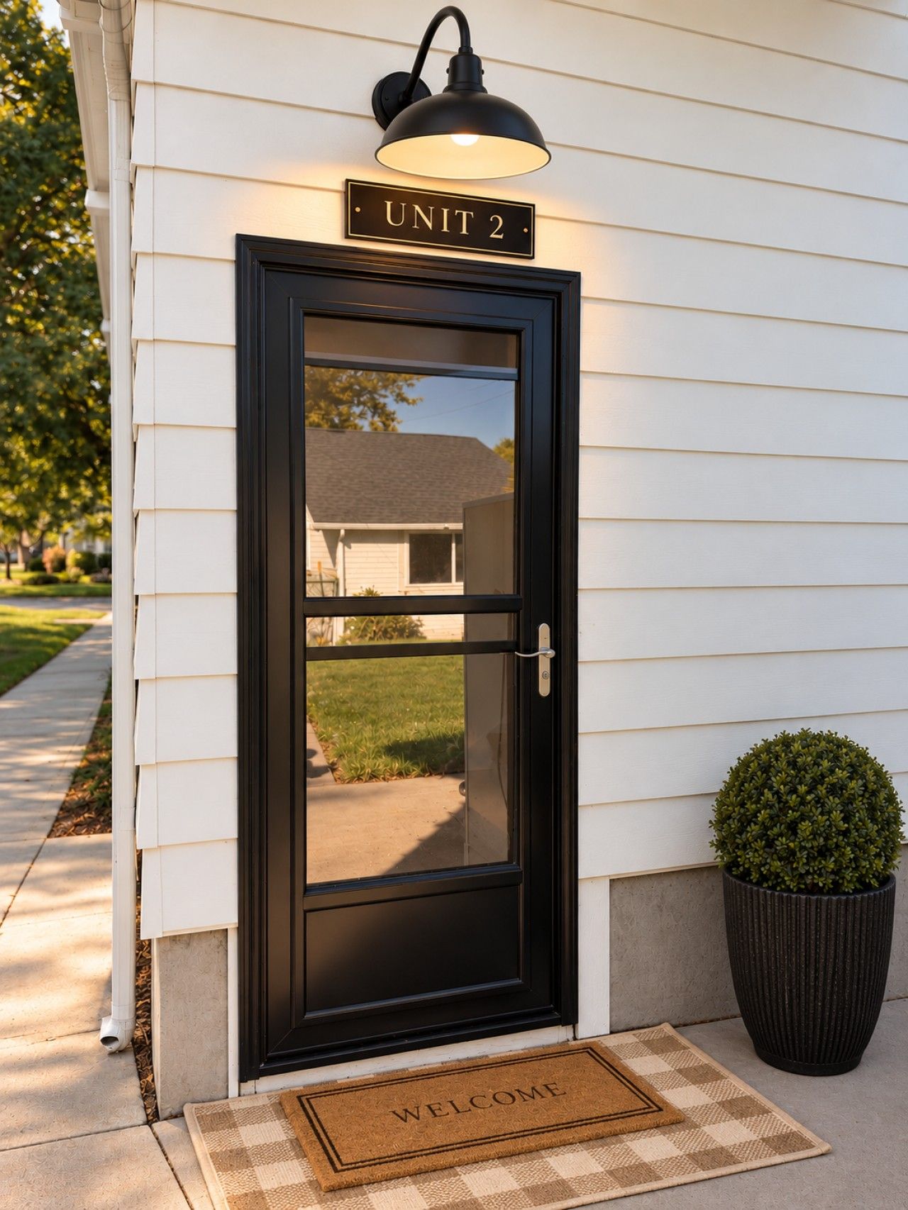 A black-framed door with a 'UNIT 2' sign above it, a small potted plant to the right, and a 'WELCOME' mat in front.