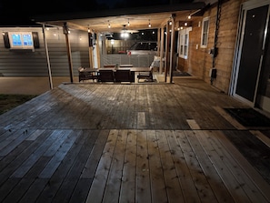 Una terraza de madera con luces colgantes, un espacio de estar techado y la entrada a un garaje.