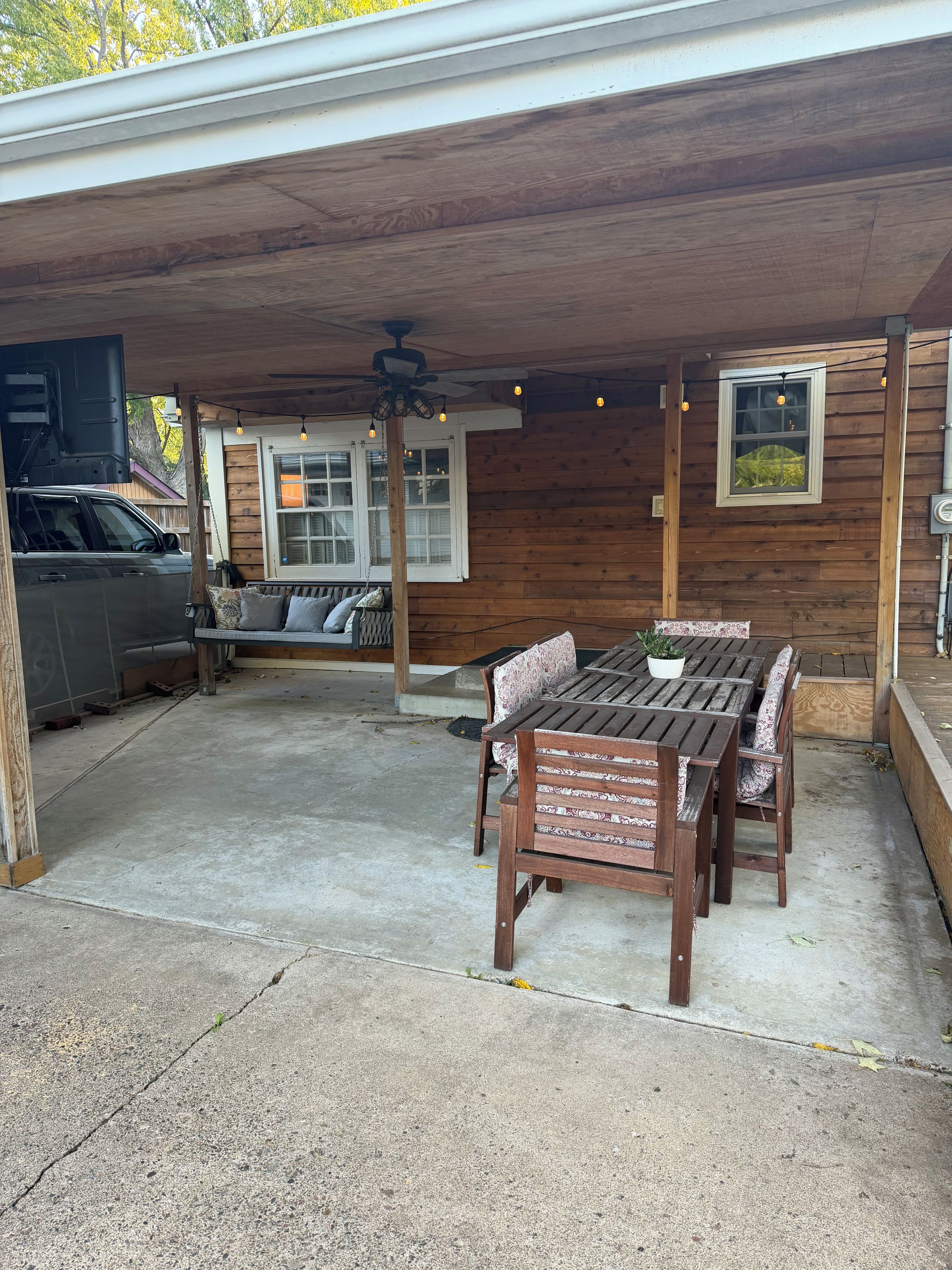 A covered patio with a wooden table and chairs, a ceiling fan, and a window with string lights.