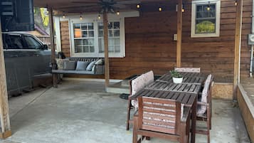 A covered patio with a wooden table and chairs, a ceiling fan, and a window with string lights.