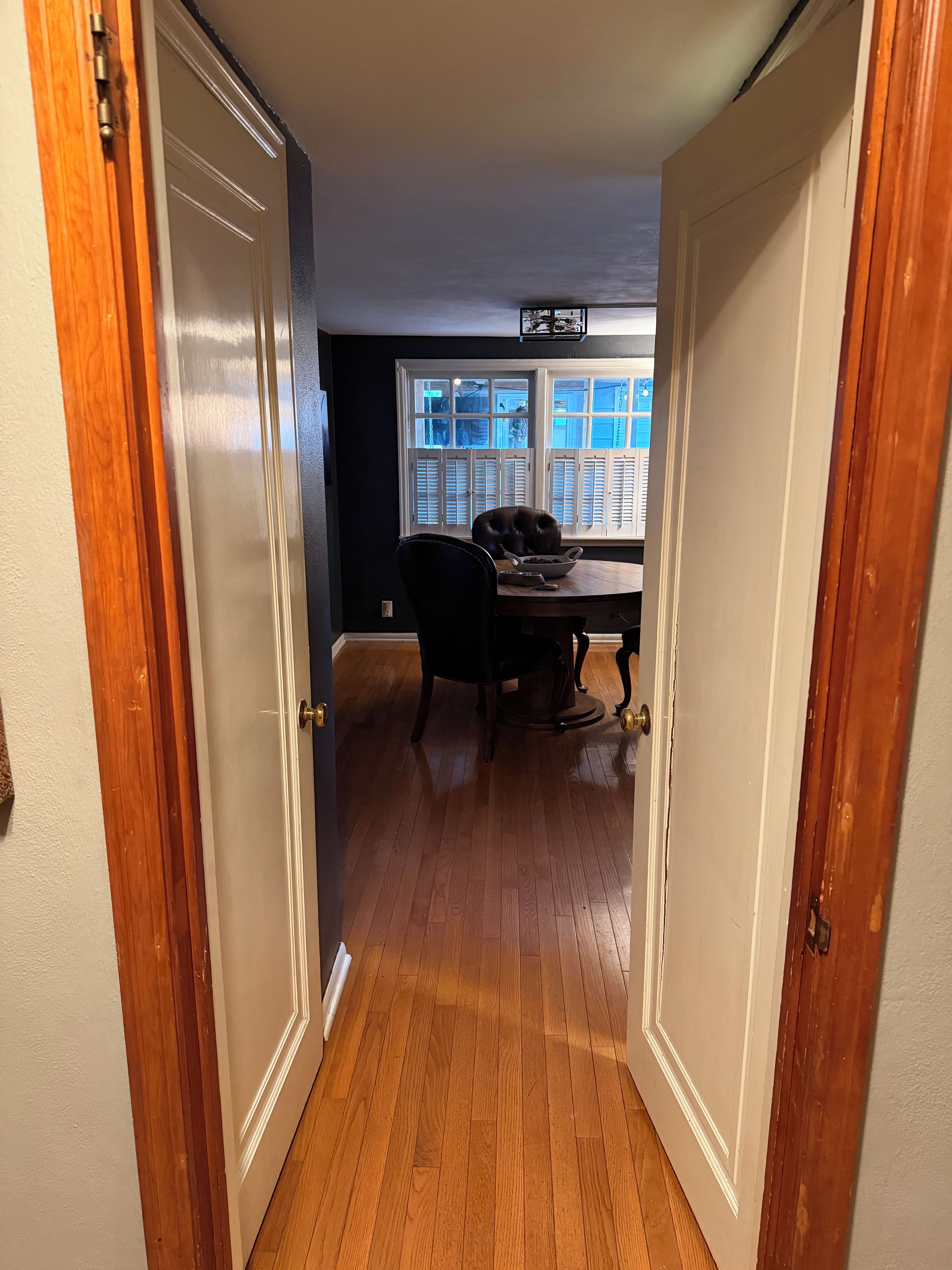 A hallway leading to a dining area with a table and chairs, a window with blinds, and a door with a brass handle.