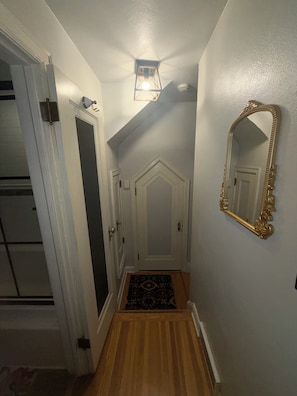 Un pasillo con piso de madera, paredes blancas, alfombra negra, puerta blanca y espejo con marco dorado.