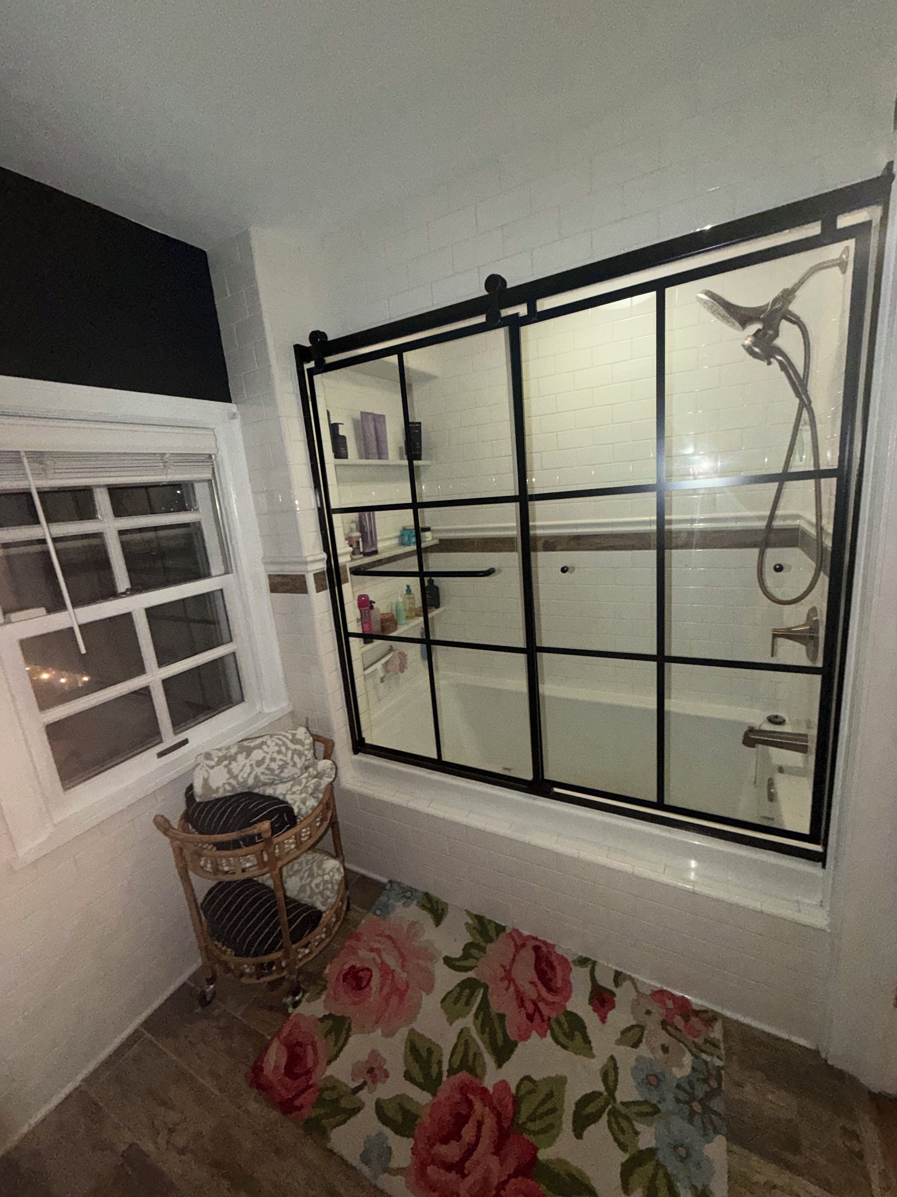 A bathroom with a bathtub, a shower, a window with a grid design, a small wicker chair, and a floral rug.