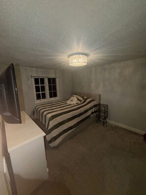 Une chambre à coucher avec un lit, un téléviseur posé sur un meuble, une fenêtre et un luminaire au plafond.