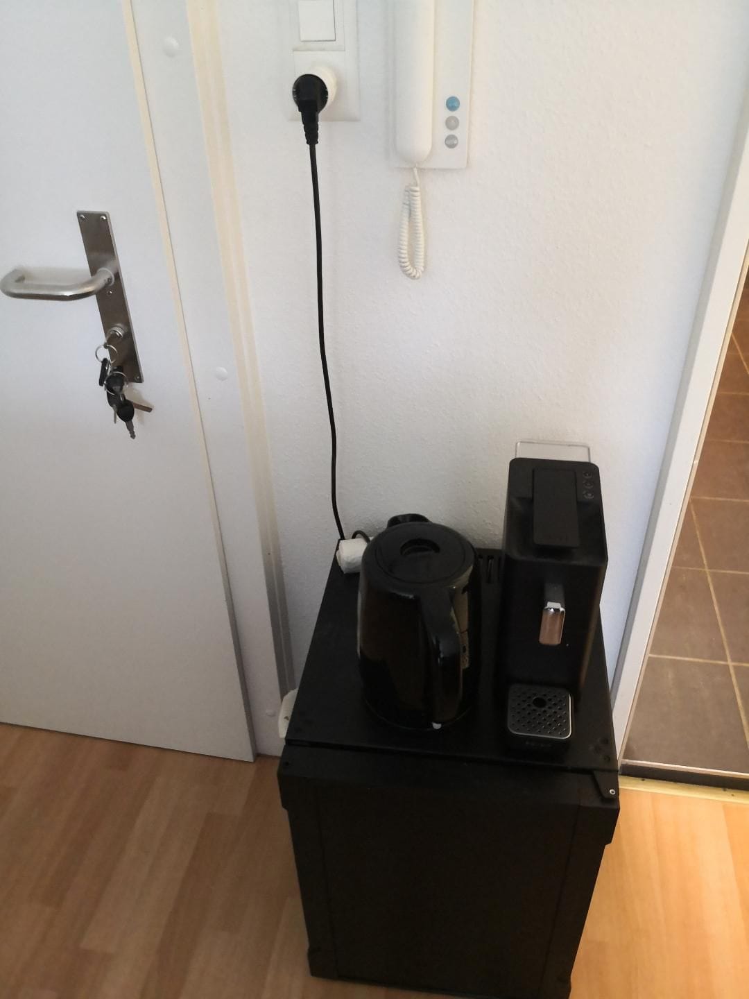 A black coffee maker on a black box, a door with a key lock, and a wall socket with a phone plugged in.