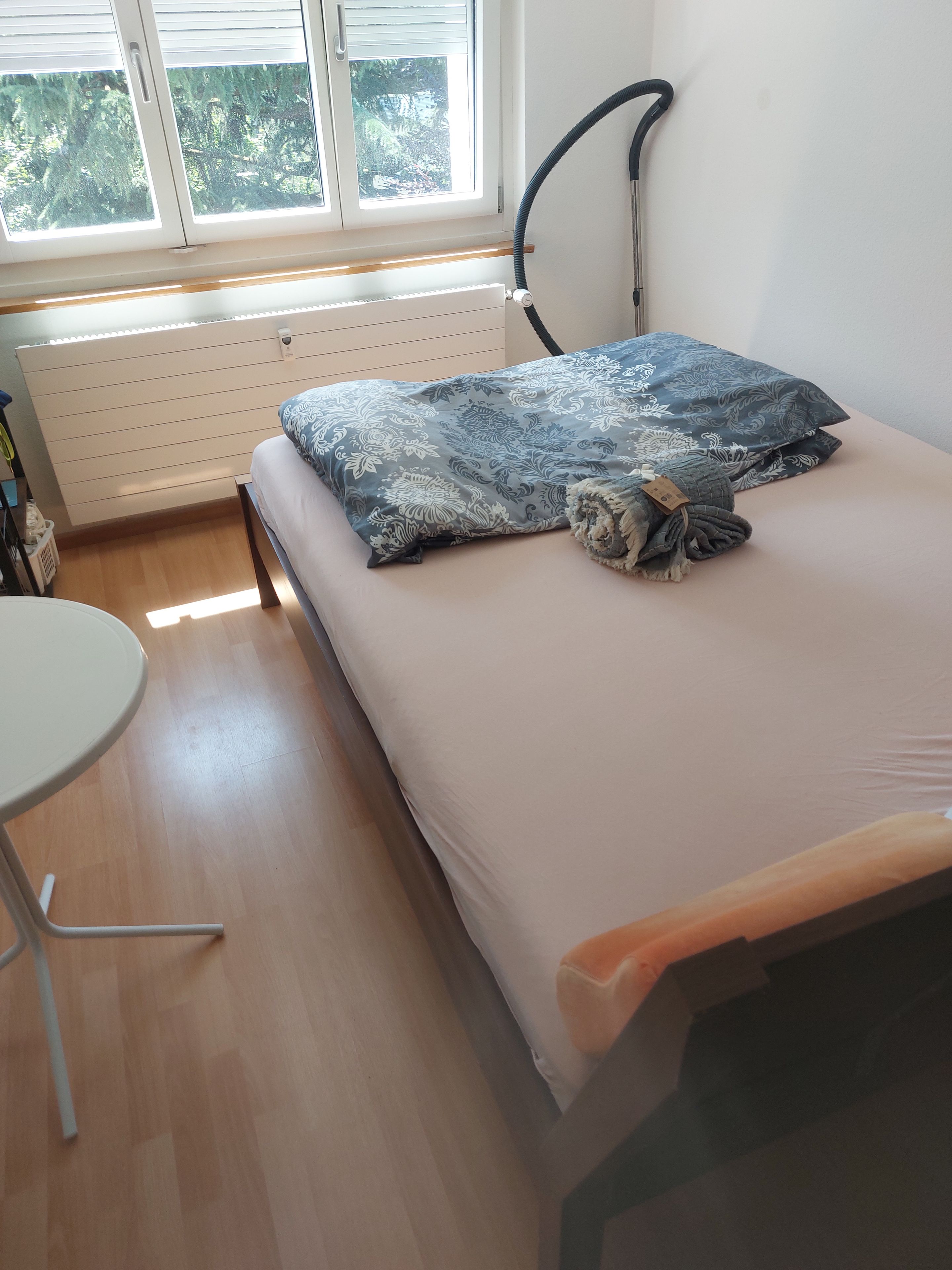 A bedroom with a bed, a radiator, a window with blinds, and a small table.