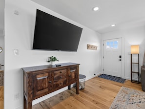 A modern living room with a flat-screen TV mounted on the wall, a wooden cabinet with drawers, a small potted plant, and a door with a window. - NEW! Ridges Retreat: Pet-Friendly Stay w/Firepit (Grand Junction)
