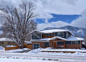 Uma casa de dois andares com telhado coberto de neve, uma grande árvore na frente e montanhas ao fundo.
