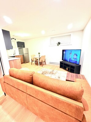 Een woonkamer met een bank, een televisie, een eethoek met stoelen, en een keuken met kasten en apparatuur.