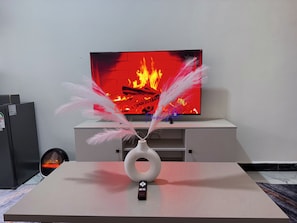Een flatscreen televisie op een witte standaard, met een decoratieve vaas en veren op een tafel ernaast.