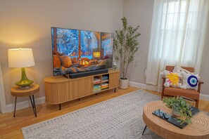 A cozy living room with a flat-screen TV, wooden entertainment unit, a green floor lamp, a potted plant, and a patterned rug. - Sunlit 2BR Near Revere Beach & Blue Line to Boston (Revere)