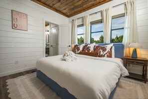 A bedroom with a large bed, a nightstand, a lamp, and a view of trees through the windows. - Mountainaire Getaway Cabin (Broken Bow)