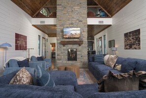 A spacious living room with a stone fireplace, blue sofas, and a large TV mounted on the wall. - Mountainaire Getaway Cabin (Broken Bow)