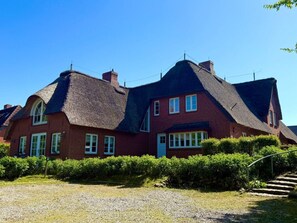Ein großes Backsteinhhaus mit einem steilen Strohdach, umgeben von gepflegten Hecken und einer Schotterauffahrt.