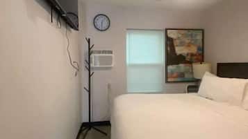 A hotel room with a bed, a clock, an air conditioning unit, a window, and a painting on the wall.