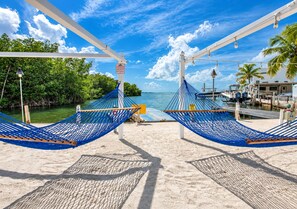A beach view with two blue hammocks under a white canopy, a clear sky, and a marina with boats. - Turtle Nest in the Keys - Cozy RV Retreat in Key Largo - Golf Cart Included (Key Largo)