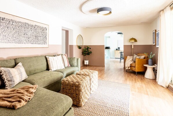 Un salon moderne avec un canapé vert, un pouf en osier, un tapis à motifs et un coin repas à l’arrière-plan.