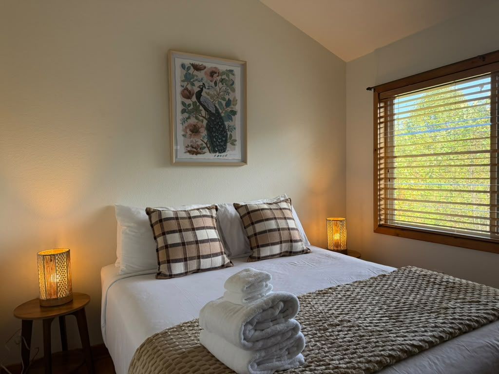Una cama bien hecha con una toalla doblada, dos almohadas, una obra de arte floral enmarcada arriba y una ventana con persianas.
