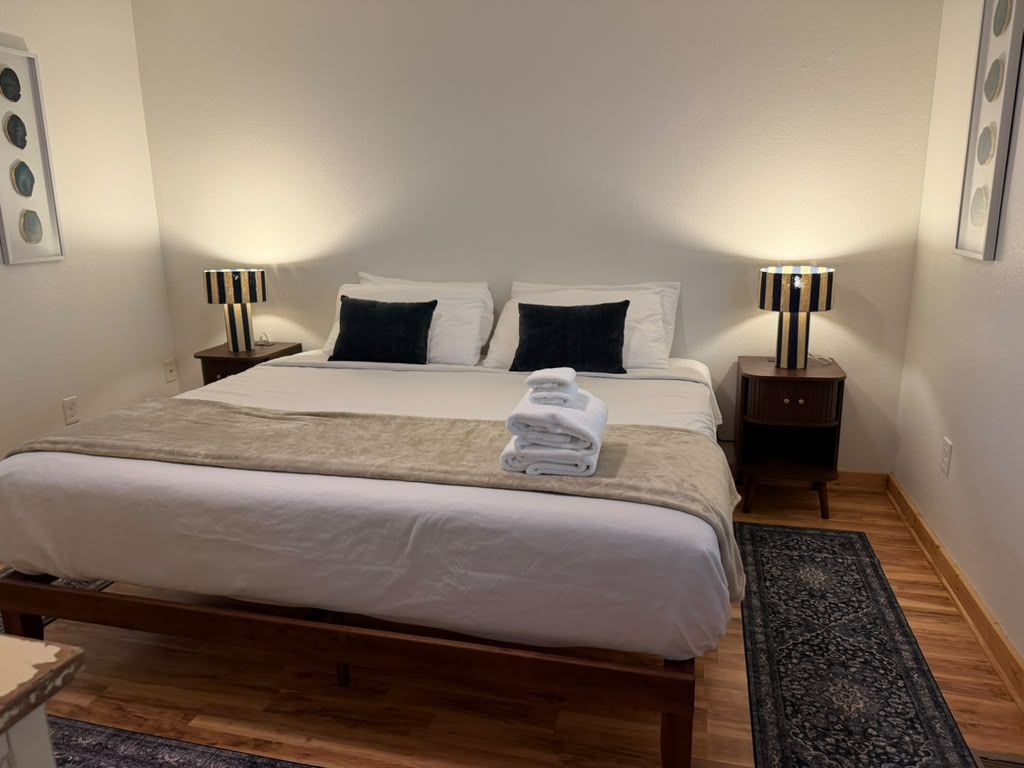Habitación de hotel con una cama grande, dos mesitas de noche con lámparas, un tapete oscuro y un cuadro colgado en la pared.