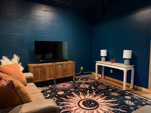 A room with a dark blue wall, a television on a wooden cabinet, a white table with lamps, and a patterned rug.