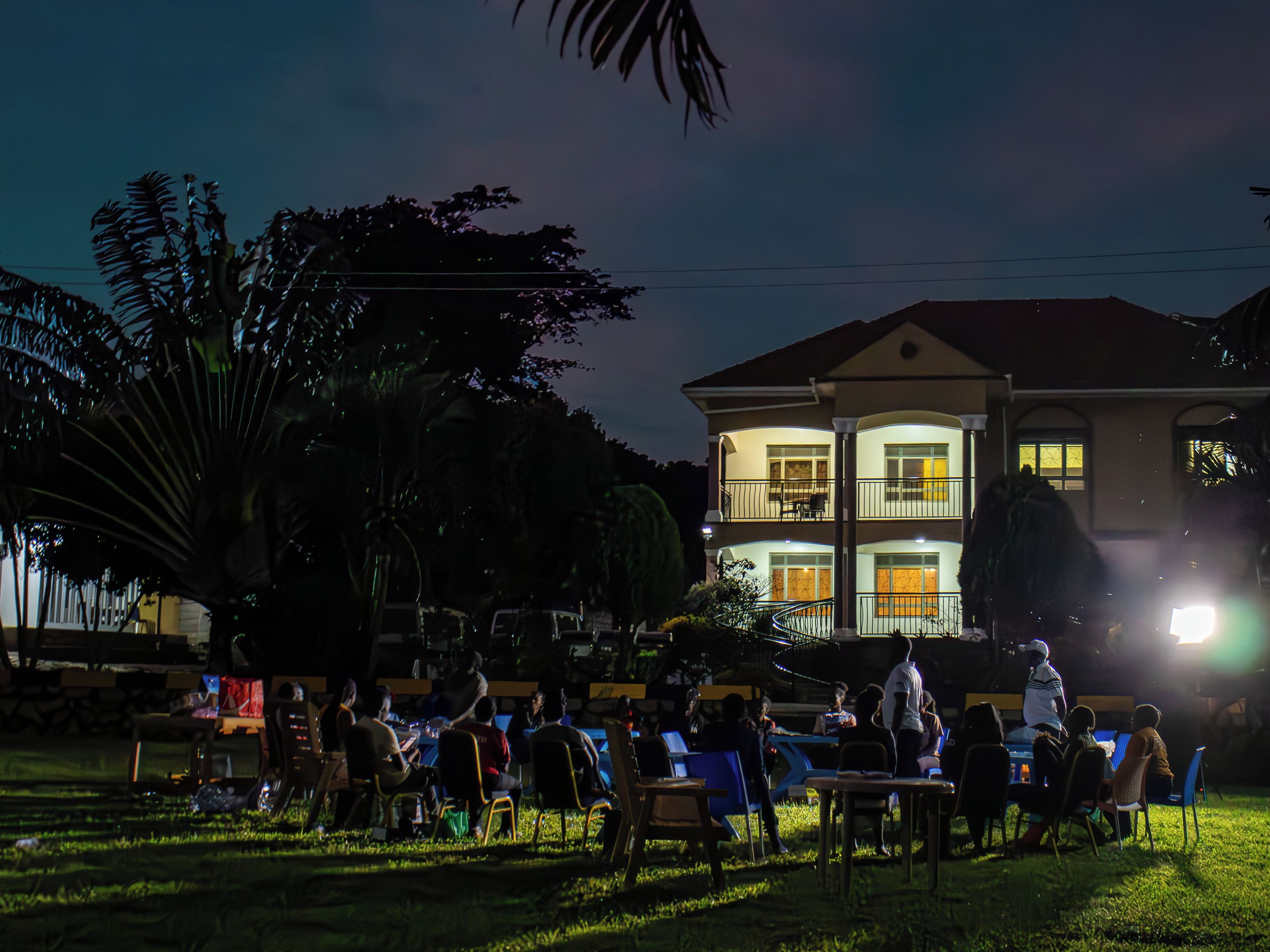 An outdoor gathering with people seated at tables, a multi-story building in the background, and a well-lit balcony.