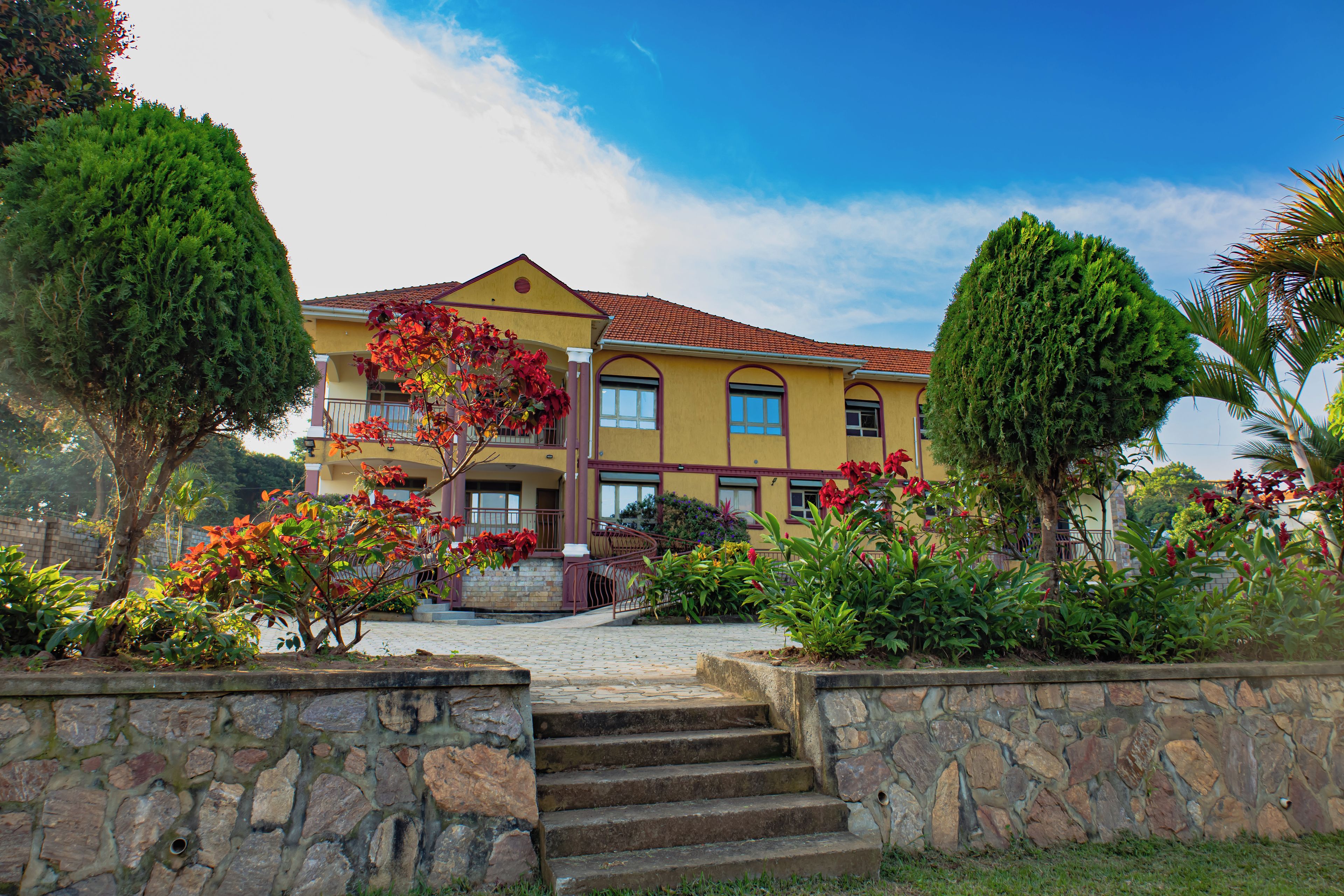 A two-story building with a stone staircase leading to the entrance, surrounded by lush greenery and flowering plants.