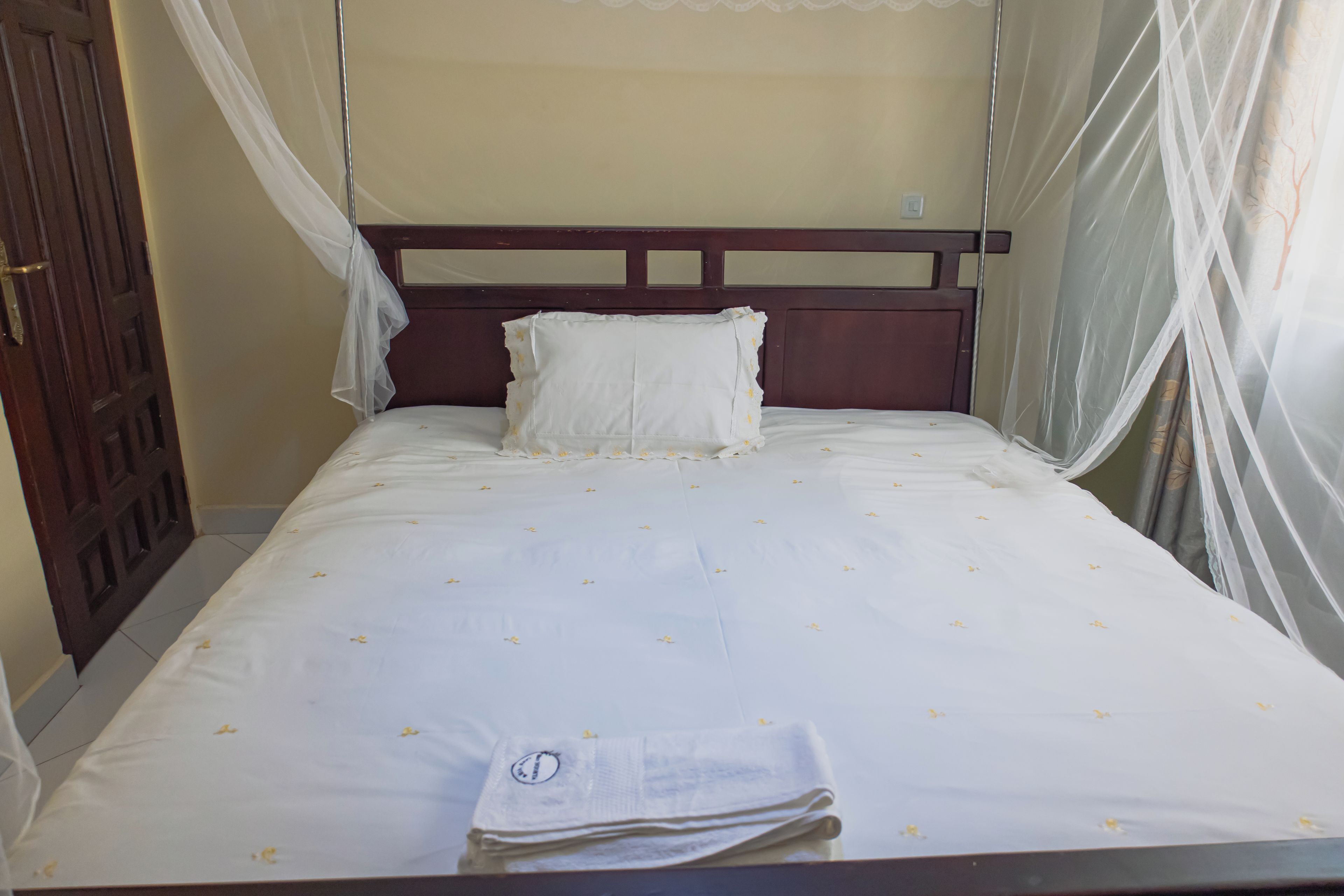 Ein Einzelbett mit weißer Bettdecke und Kissen, ein hölzernes Kopfholz und ein Fenster mit durchsichtigen Vorhängen.