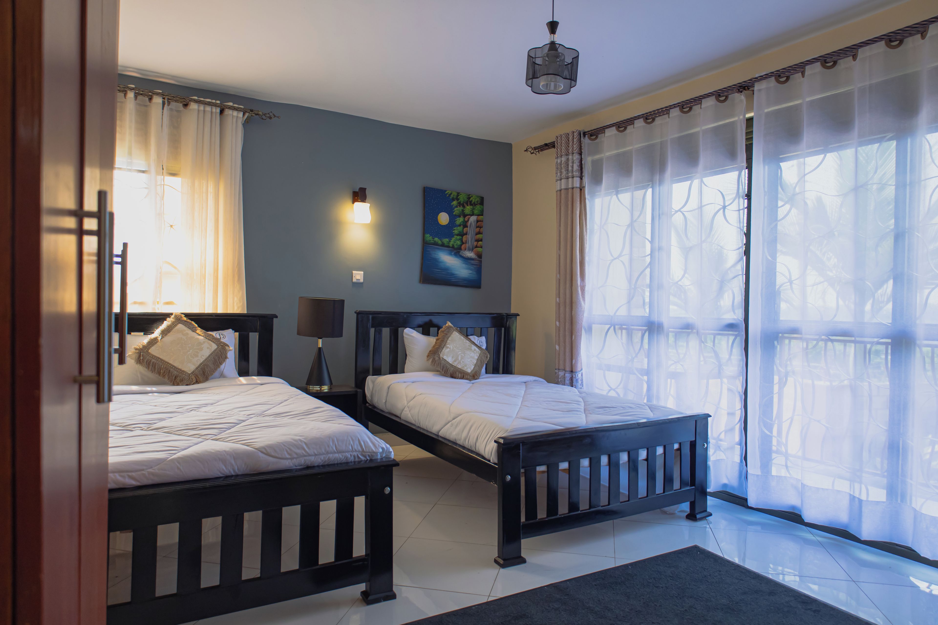 Un dormitorio con dos camas, una mesita de noche, una lámpara, un cuadro y una ventana con cortinas.