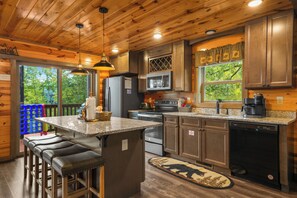 A modern kitchen with wooden cabinets, a central island with bar stools, stainless steel appliances, and a view of the outdoors through large windows. - Sleeps 12, Private Indoor Pool, Hot Tub, Theater, Arcade, 6 Mi to Dollywood (Pigeon Forge)