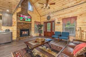 A log cabin interior with a fireplace, a television mounted on the wall, a dining table, and a seating area with chairs. - Sleeps 12, Private Indoor Pool, Hot Tub, Theater, Arcade, 6 Mi to Dollywood (Pigeon Forge)