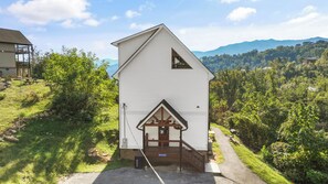 A white building with a triangular roof and a small porch, surrounded by greenery and mountains in the background. - Sleeps 10, King Bed in Every Room, Hot Tub, Views, 3 Mi to Downtown Gatlinburg (Gatlinburg)
